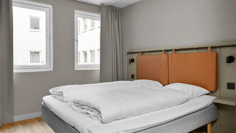 Et hyggeligt hotelværelse med en komfortabel seng, hvidt sengetøj og to vinduer, der slipper naturligt lys ind. Perfekt til et afslappende ophold.