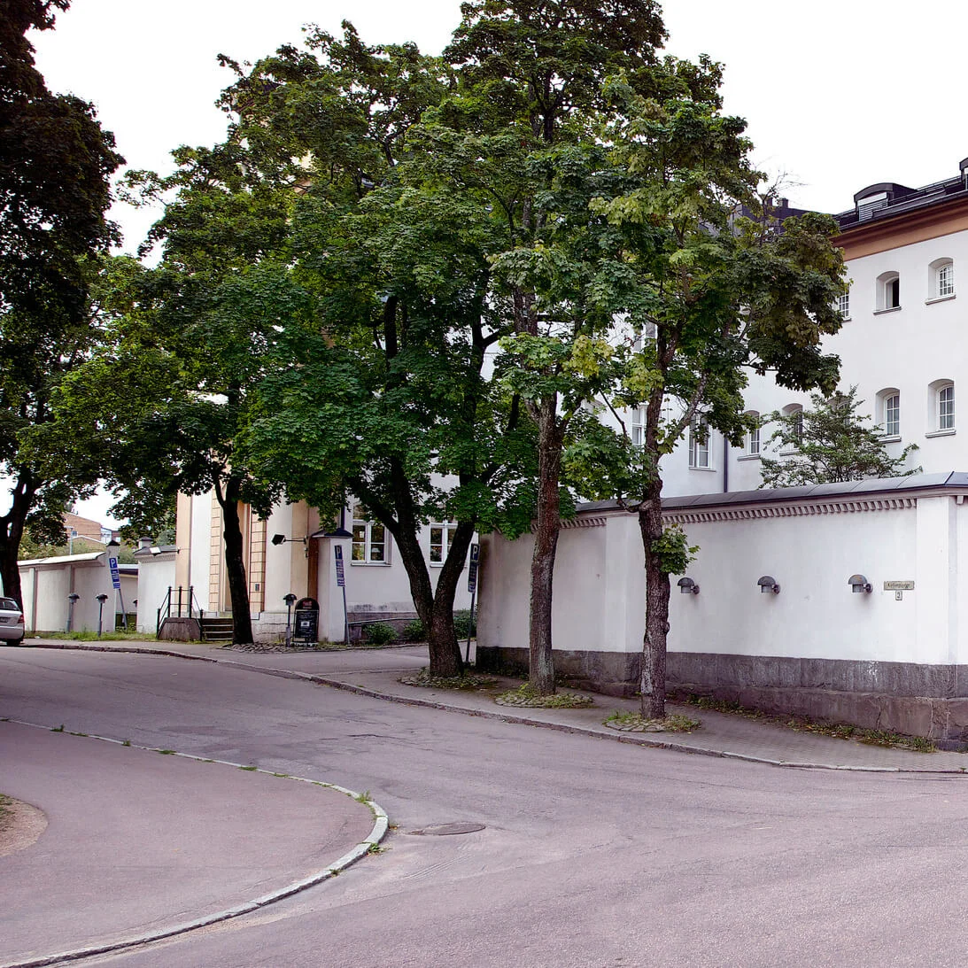 A historic building at a street corner, surrounded by a high wall, with a tree-lined sidewalk and multiple national flags flying.