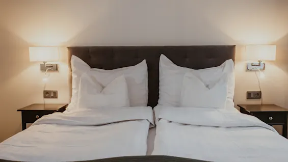 Et hyggeligt hotelværelse med to senge, hvidt sengetøj og varm belysning fra vægmonterede lamper, der antyder komfort og afslapning.