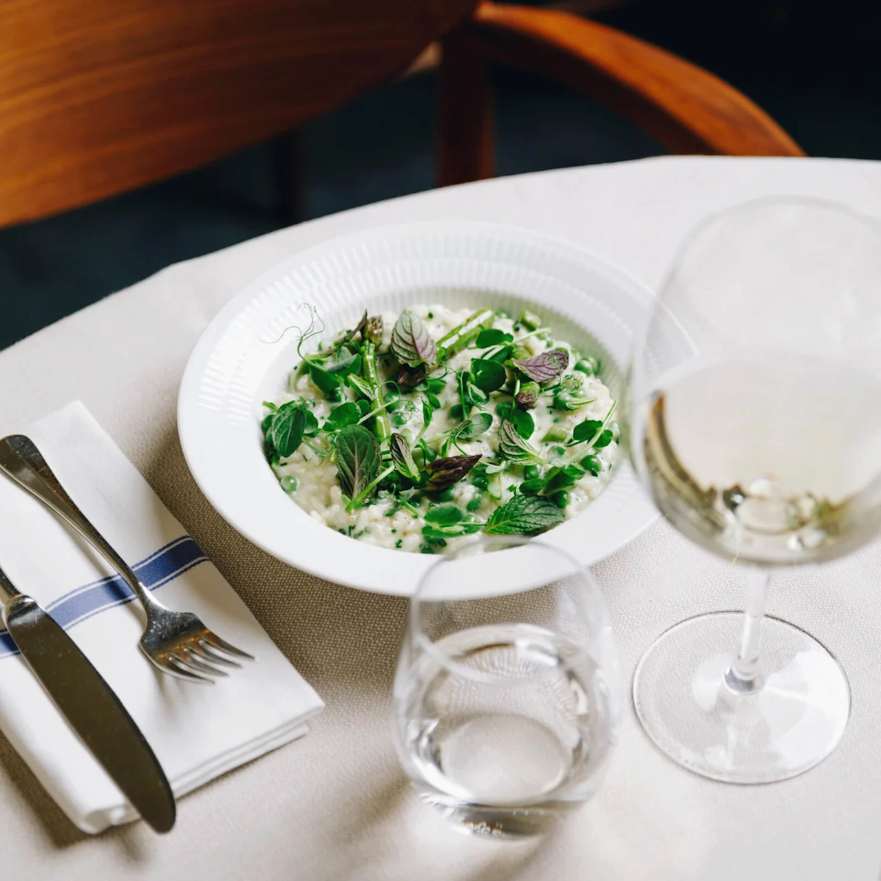 A plate of risotto topped with green herbs sits on a white tablecloth beside a glass of white wine, cutlery on a napkin, and a wooden chair.