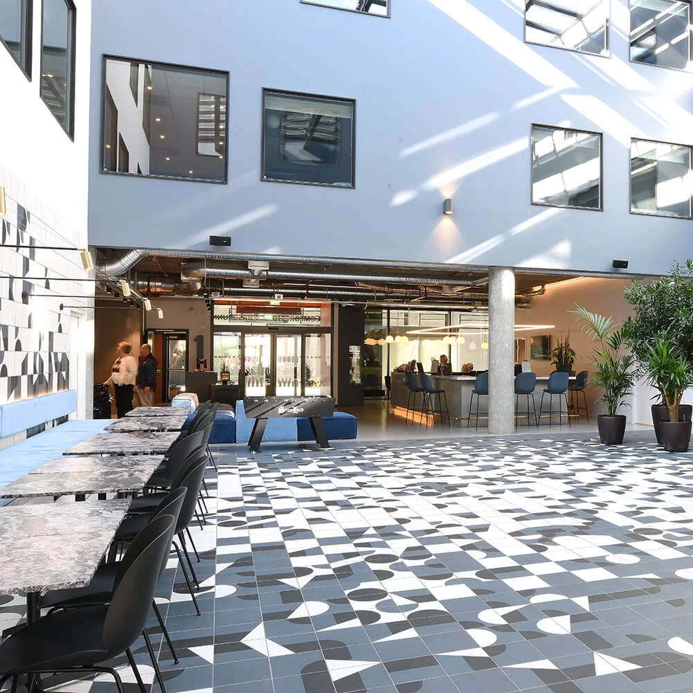 Farverige geometriske fliser dækker gulvet i et moderne atrium med borde, stole og potteplanter. Naturligt lys strømmer ind gennem flere vinduer og skaber en lys, luftig atmosfære.