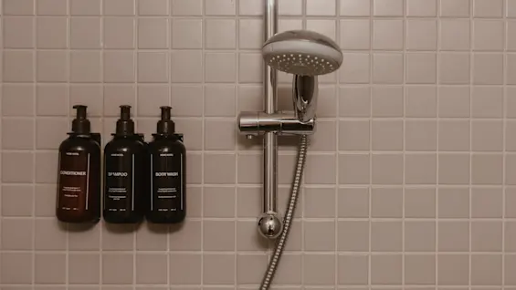 Suihkupää kiinnitetty kaakeloituun seinään kiiltävän metallisen ohjauspaneelin yläpuolelle. Kolme pulloa, joissa on merkinnät "CONDITIONER", "SHAMPOO" ja "BODY WASH", on siististi sijoitettu vasemmalle.