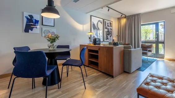 A modern dining area with blue chairs, a round table, artwork on walls, and a wooden sideboard in a well-lit room with a balcony door.