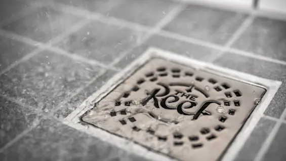A metal drain cover with text "The Reef" collects splashing water on a tiled floor, indicating a wet environment.