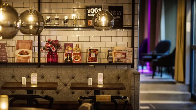 En koselig kaféinteriør med stoppet benkesete, hengende lys, og dekorative veggfliser som viser plakater og aviser.