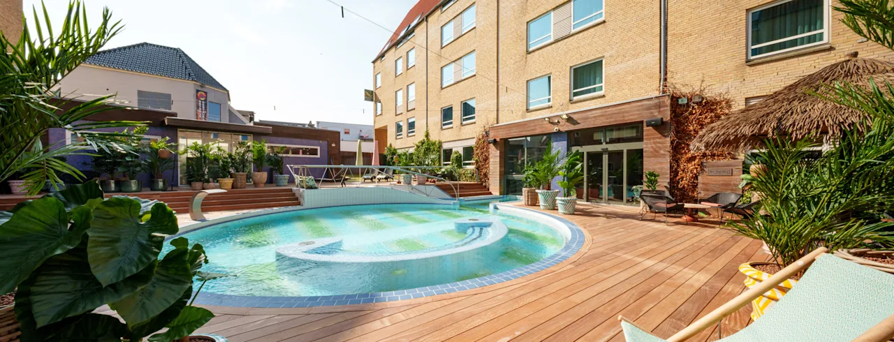 Pyöreä uima-allas, ympäröitynä puisella terassilla, aurinkotuoleilla ja ruukkukasveilla, vieressä monikerroksinen tiilirakennus suurine ikkunoineen.