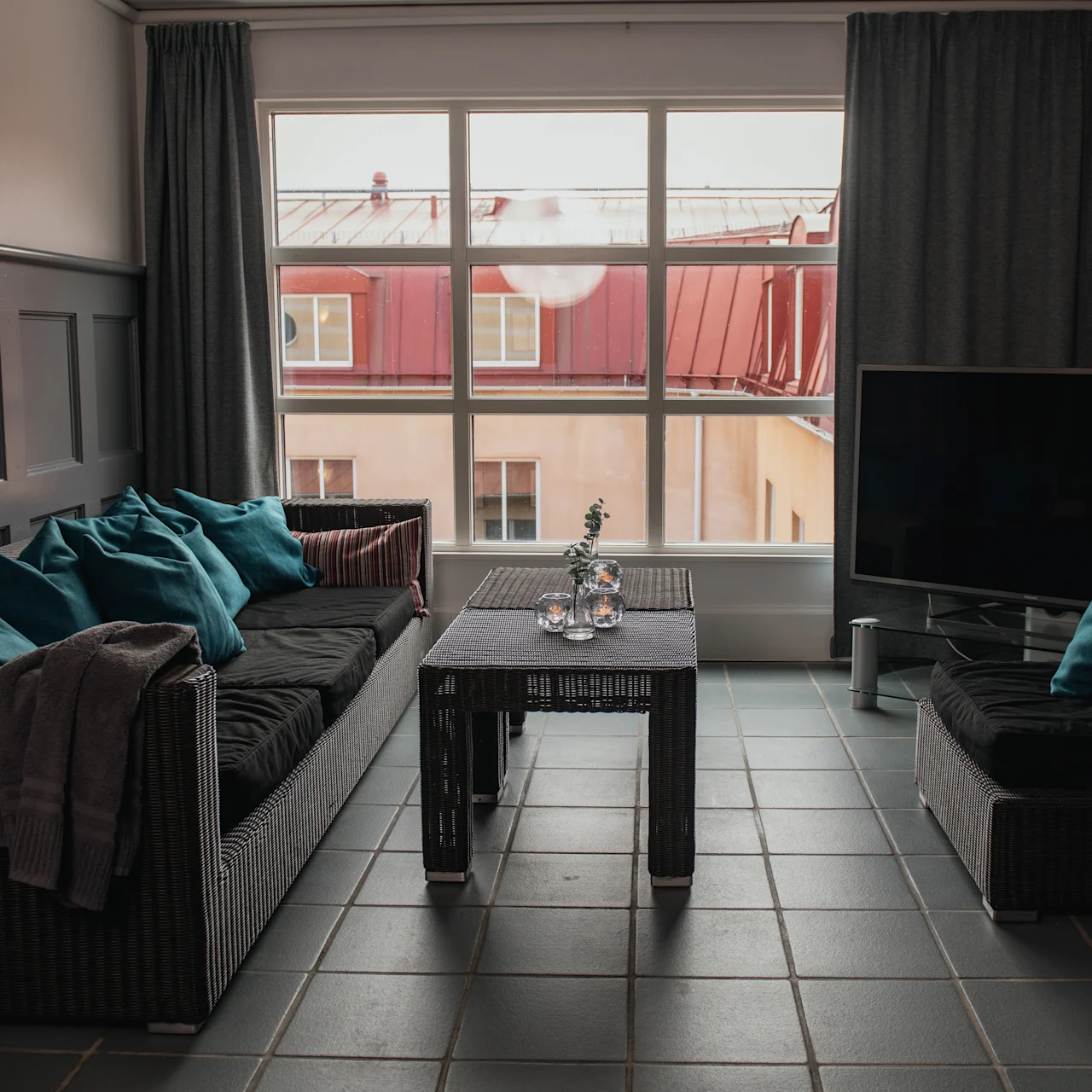 A living room with a wicker sofa set adorned with teal pillows surrounds a table with vases. Large windows reveal a rooftop view. A flat-screen TV is on the right.