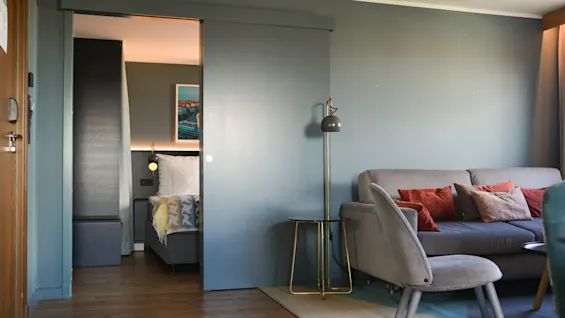 En grå sofa i stuen med farverige puder står ved siden af en moderne gulvlampe. En skydedør afslører delvist et soveværelse med en seng og vægkunst.
