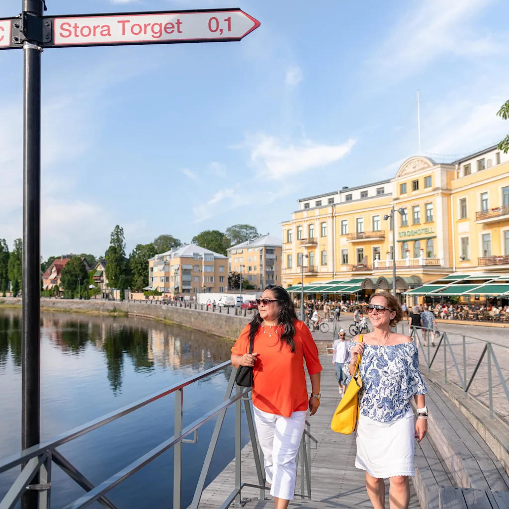 Two women walking along a sunny waterfront boardwalk next to a sign pointing towards Stora Torget