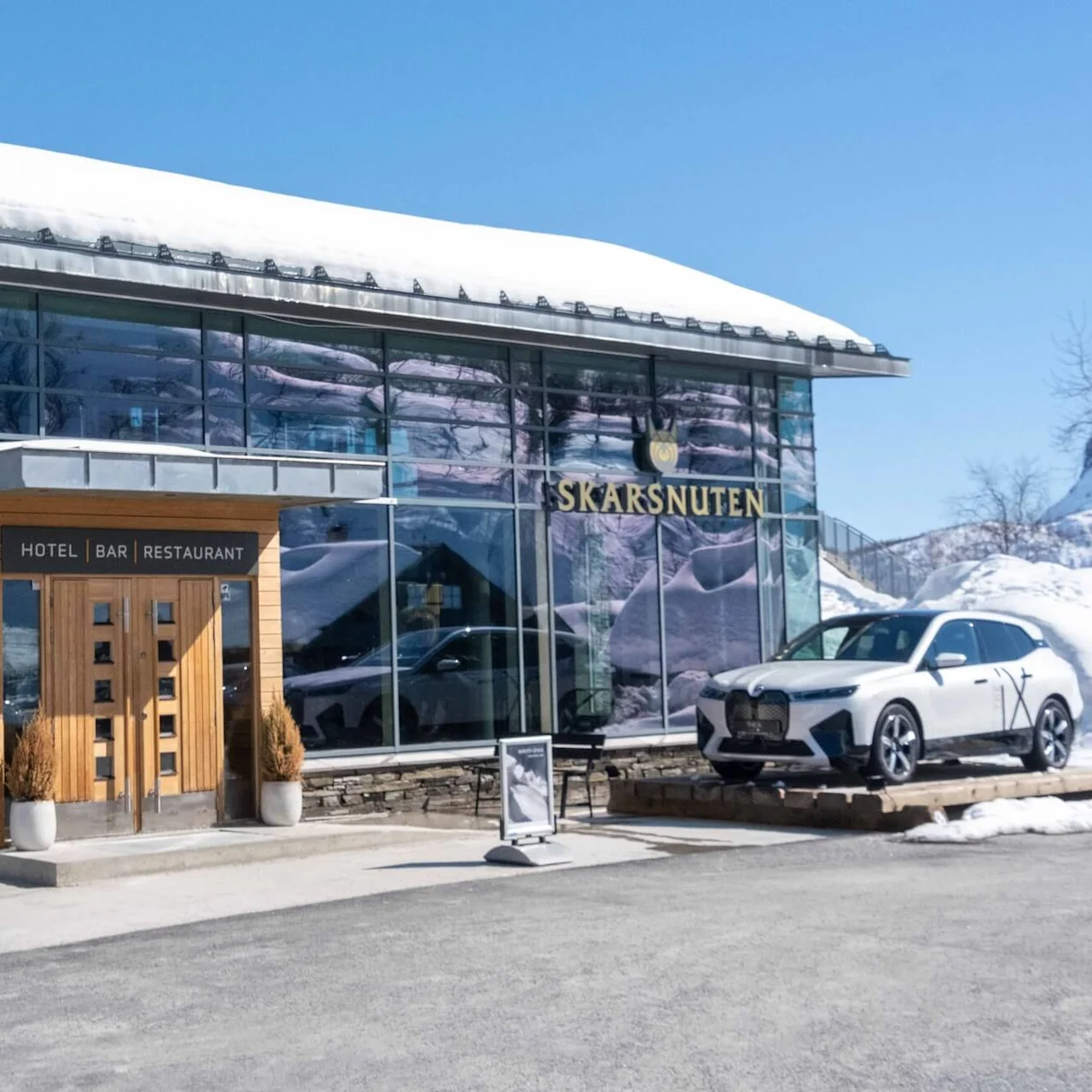 A skier in orange gear walks with skis on shoulders outside a snowy hotel with signage: "SKARSNUTEN", "HOTEL | BAR | RESTAURANT". A white car is parked nearby under clear blue skies.