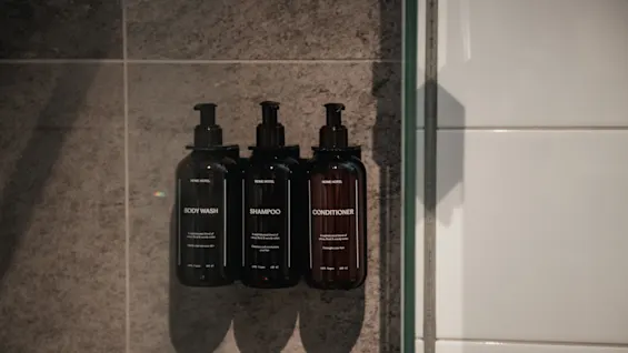 Tre mørke flasker merket Body Wash, Sjampo og Balsam montert på en flislagt dusjvegg, klare til bruk.