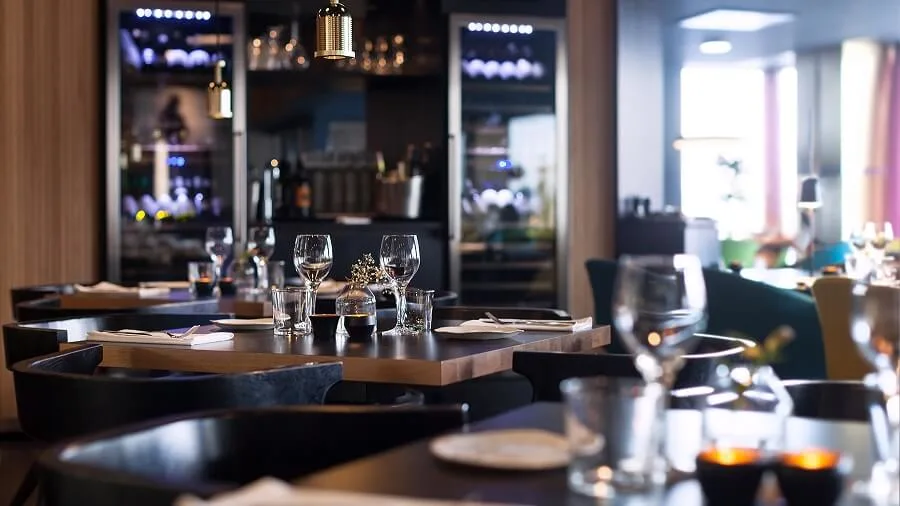 Borde dækket med glas og servietter i en hyggelig, blødt belyst restaurant, omgivet af trævægge og vinudstillinger, hvilket skaber en intim spiseatmosfære.