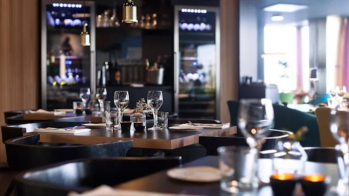 Bord dekket med glass og servietter i en koselig, mykt opplyst restaurant, omgitt av trevegger og vinutstillinger, som skaper en intim spisestemning.