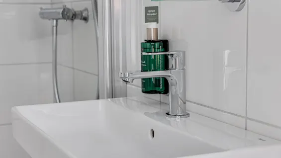 A white bathroom sink with a silver faucet in a tiled setting; a green bottle labeled "shower gel" and "shampoo” is placed beside it.