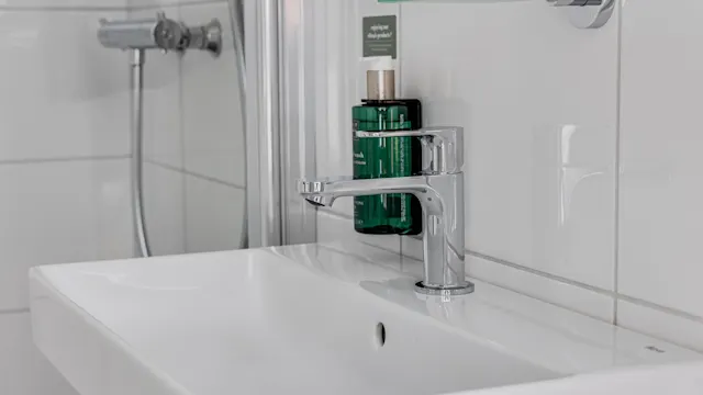 Valkoinen kylpyhuoneen lavuaari, jossa on hopeinen hana laatoitetussa ympäristössä; vieressä vihreä pullo, jossa lukee "suihkugeeli" ja "shampoo".