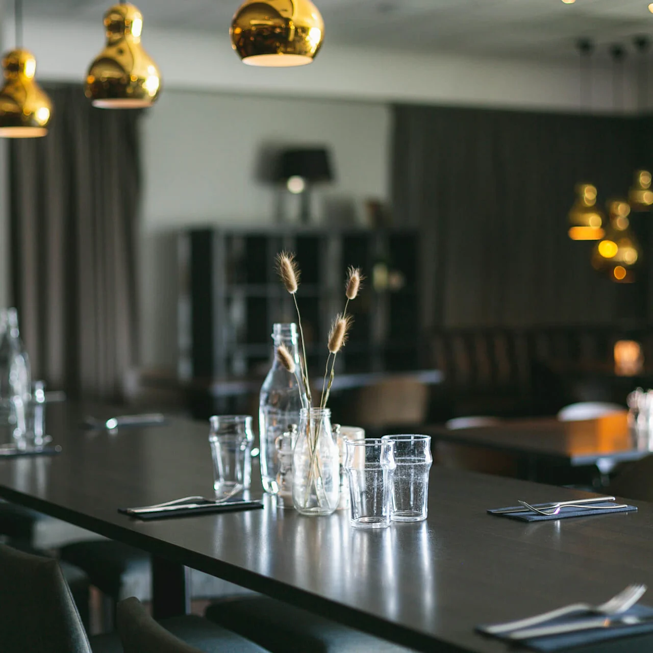 Elegant spisebord dækket med glas, bestik og en krukke med tørrede blomster, under varme, lysende pendellamper i en svagt oplyst restaurant.