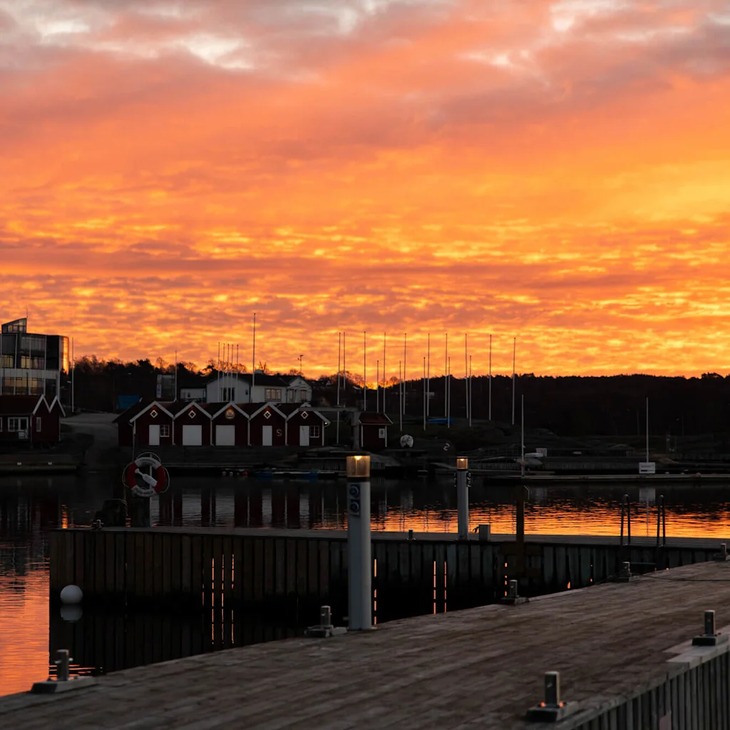 Träpiren reflekterar en livlig orange solnedgång; små röda bodar och en modern glasbyggnad i bakgrunden; stilla vatten omger, silhuett av master mot färgglad himmel.