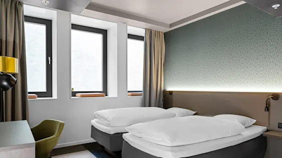 Two neatly made single beds stand side by side in a modern room with green wallpaper, a desk with a chair, and large windows partially covered by curtains.
