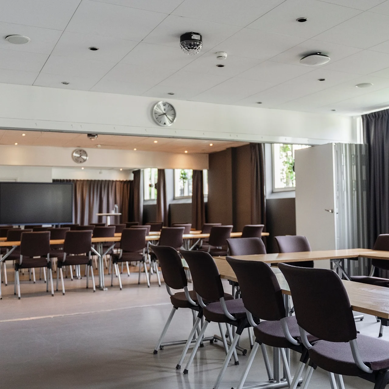 Konferensrum med rader av bruna stolar och bord, arrangerade mot en stor skärm. Utrymmet är välbelyst av fönster med växter, under en takklocka och lampor.