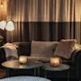 Et hyggeligt loungeområde med en mørk sofa, to runde borde med glødende stearinlys og varm belysning fra gulvlamper. KOMNA!