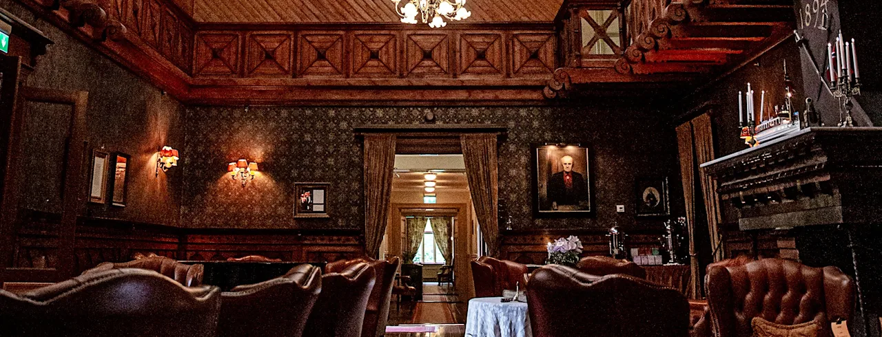 Grand chandelier hanging from an ornate stained glass ceiling in a luxurious, wood-paneled room with elegant furnishings, framed portraits, and a large fireplace. 