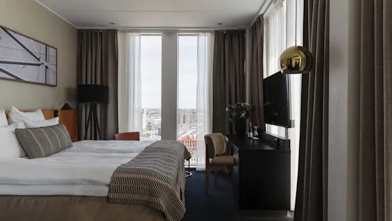 Et moderne hotellrom med en komfortabel seng, store vinduer med utsikt over byen og et skrivebord med TV.
