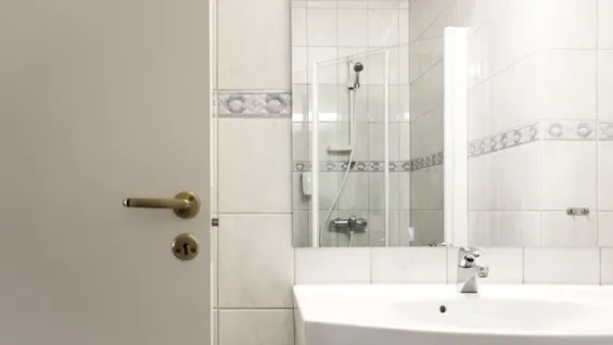Door with brass handle, closed; behind it, a bathroom with white tiles and blue patterns. Visible are a sink with a faucet and a glass shower enclosure.