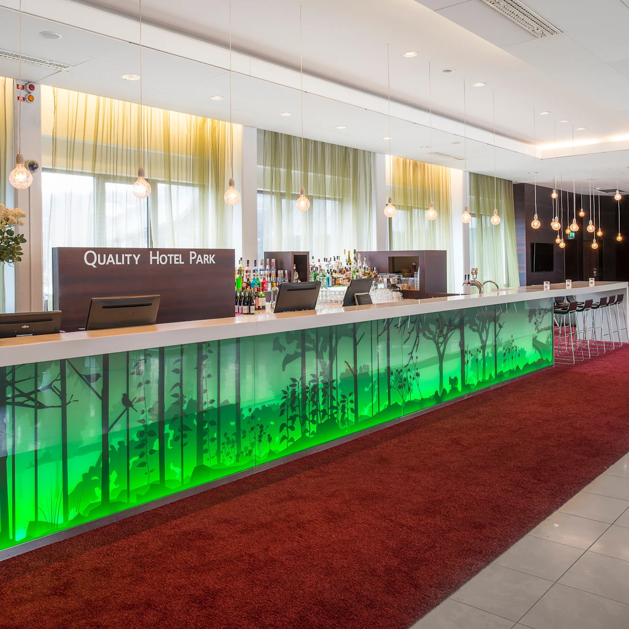 Resepsjonsdisk med grønne skogbilder, opplyst og dekorert, plassert på rød teppe i en moderne hotellobby. Teksten lyder "QUALITY HOTEL PARK."