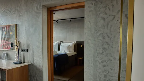 Ett modernt sovrum med en prydligt bäddad säng i bakgrunden. En grå texturerad vägg har en dörröppning med träram. Ett litet skrivbord med en lampa och vas står till vänster.