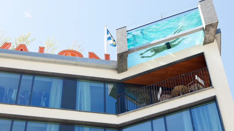 A rooftop pool with a swimmer above a modern building facade, featuring glass windows and a balcony with seating. The sky is clear. Text: "KAUAI PALACE HOTEL."