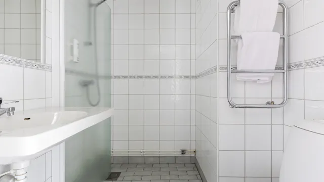 Ett vitt, kaklat badrum med handfat och spegel till vänster, en handdukshängare med vikta handdukar till höger, och en dusch i bakgrunden.
