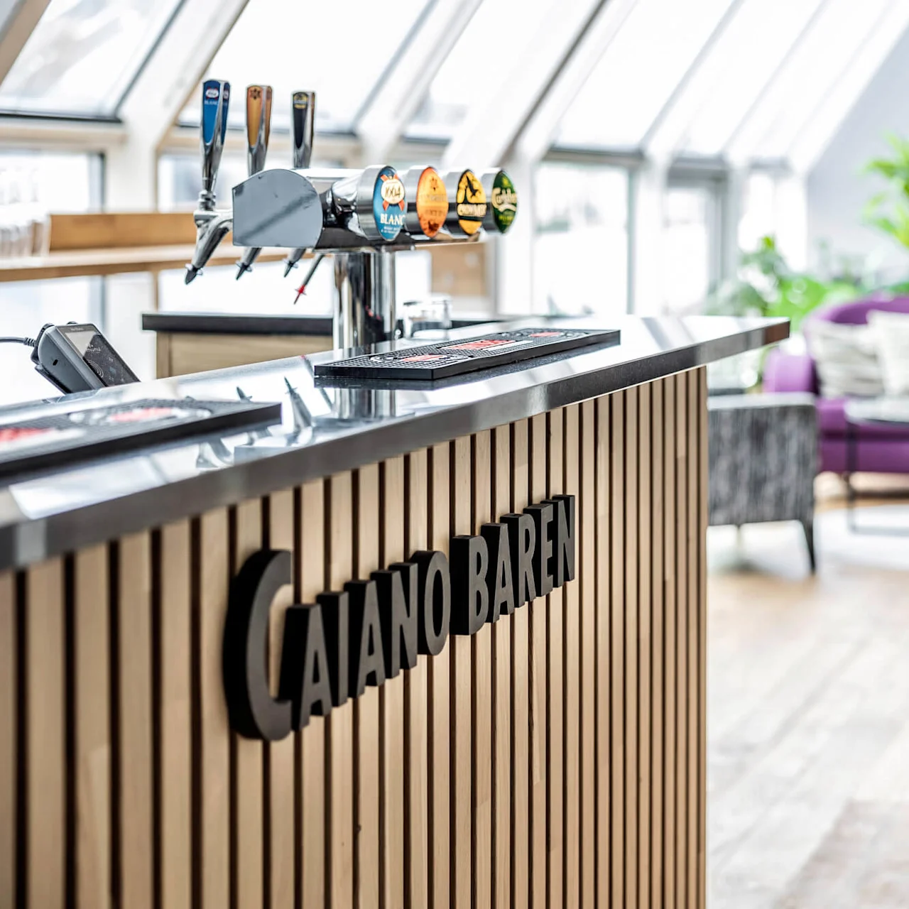 Barräknaren har öltappar och kortterminal; texten "CAIANO BAREN" på framsidan. Modern lounge med lila soffa, grå stolar och abstrakt konst i bakgrunden.
