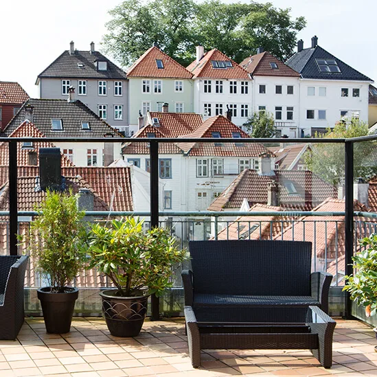 Udendørs siddeområde med sorte fletmøbler og potteplanter på en flisebelagt terrasse, med udsigt over en charmerende udsigt af traditionelle huse med varierende tagdesign.