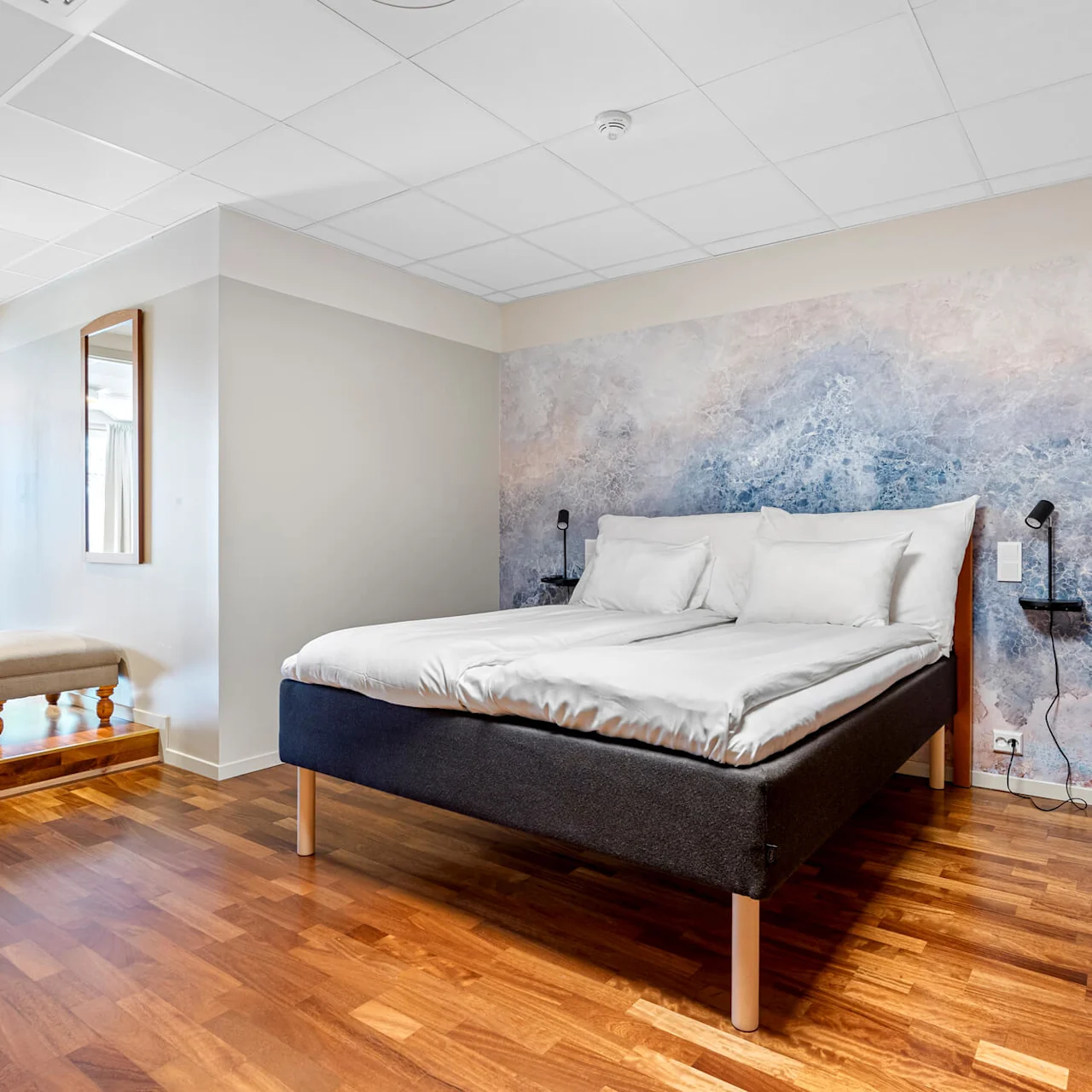 A bed with white bedding is centered against an abstract blue wall mural in a bright bedroom with wood flooring.