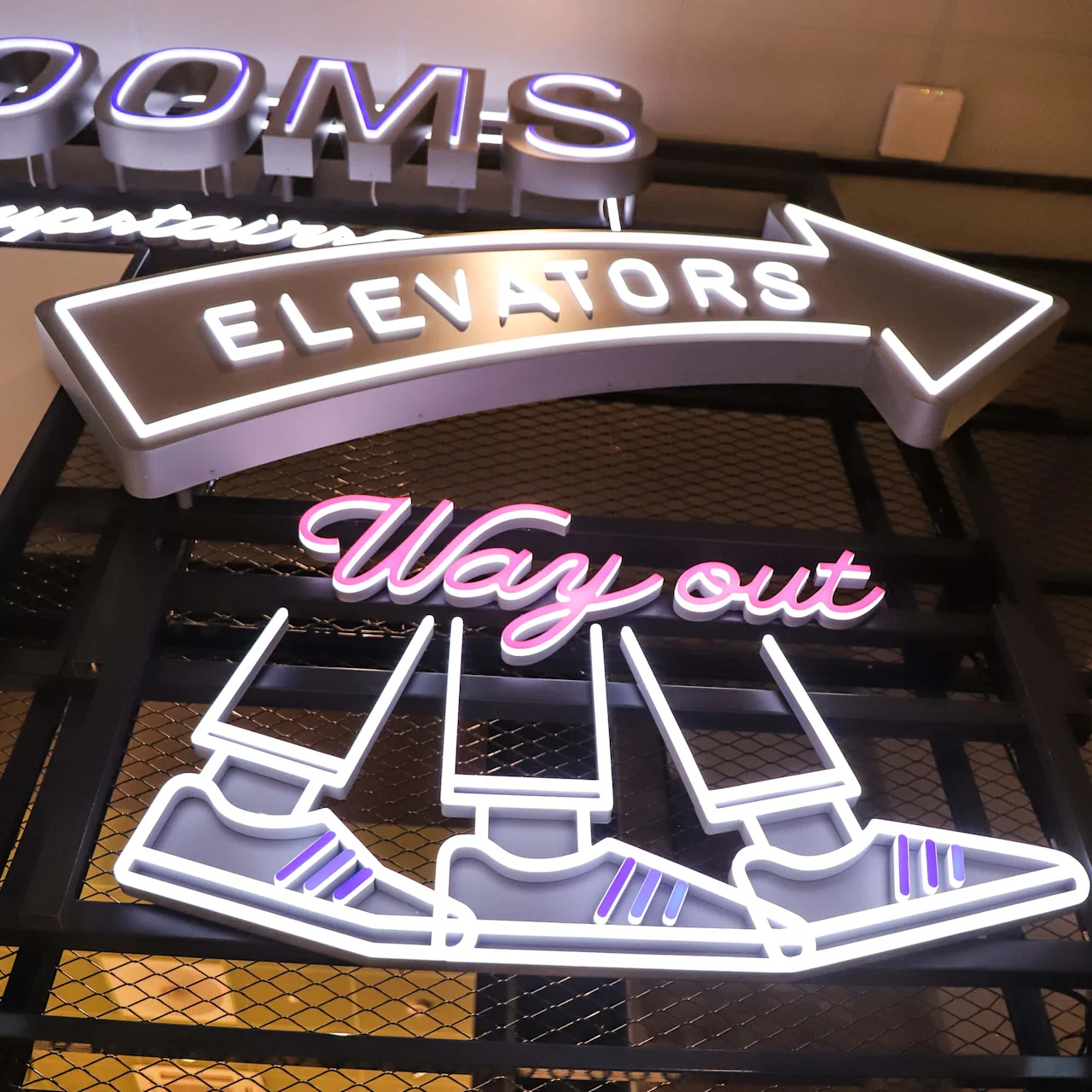 Neonskilt viser "ELEVATORS" i en pil, over "Way out" og sko grafikk, montert på et nett bakgrunn. Delvis tekst "ROOMS" synlig ovenfor.