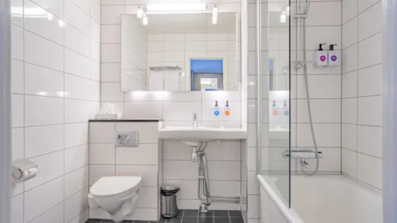 Toalett, servant og badekar/dusj i et moderne, hvitflislagt bad med et stort speil, under sterkt lys. Sjampo- og dusjsåpedispensere er montert på veggen.