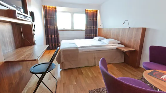 A neatly made bed rests against a wooden headboard, accompanied by a small bedside table in a hotel room. Nearby, purple chairs and a small table complete the cozy setting.