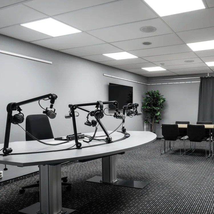 En modern podcaststudio med mikrofoner och hörlurar på ett centralt bord. En rad stolar och en TV-skärm finns i bakgrunden. Skylten lyder "NÖDUTGÅNG".