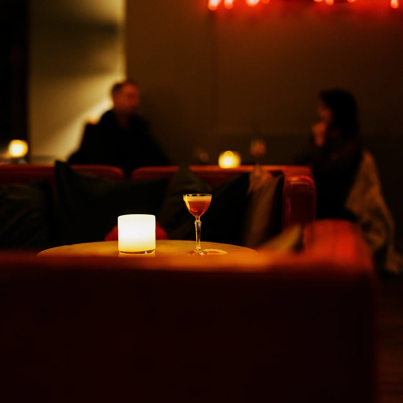 A cocktail glass sits on a table beside a lit candle, in a dimly lit bar with neon text on the wall reading "Wine a bit," two blurred figures converse in the background.