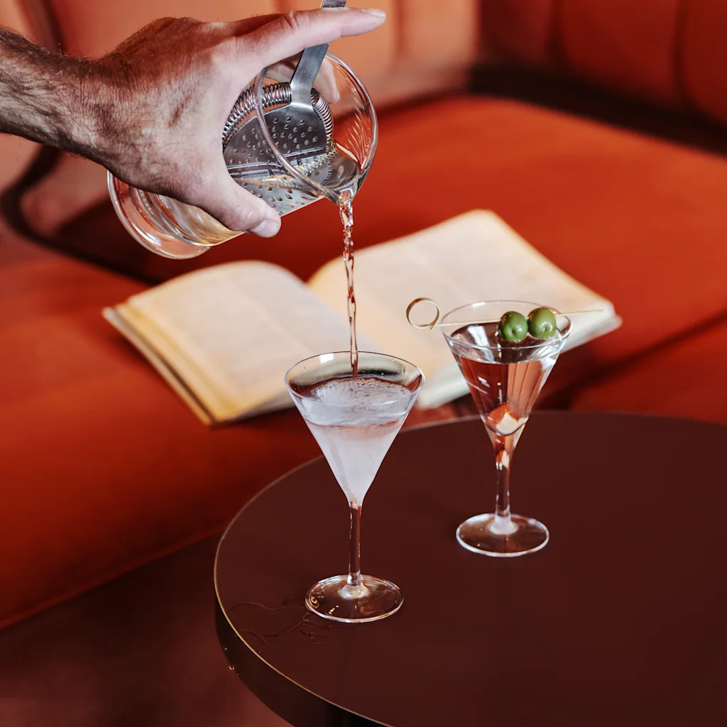 A hand pours a cocktail into a martini glass on a table next to another drink and an open book on a red sofa.