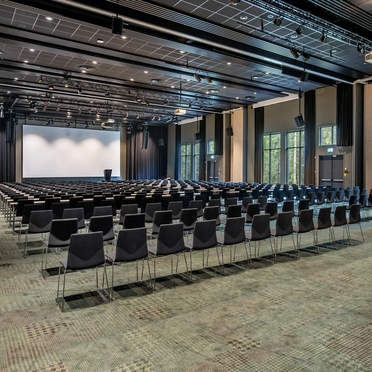 Rows of empty chairs face a large projection screen in a spacious auditorium with tall windows showing a forest view.