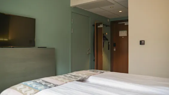 En seng med hvide lagener og et mønstret tæppe er placeret i et hotelværelse. Et TV er monteret på en væg overfor. Rummet inkluderer en trædør.