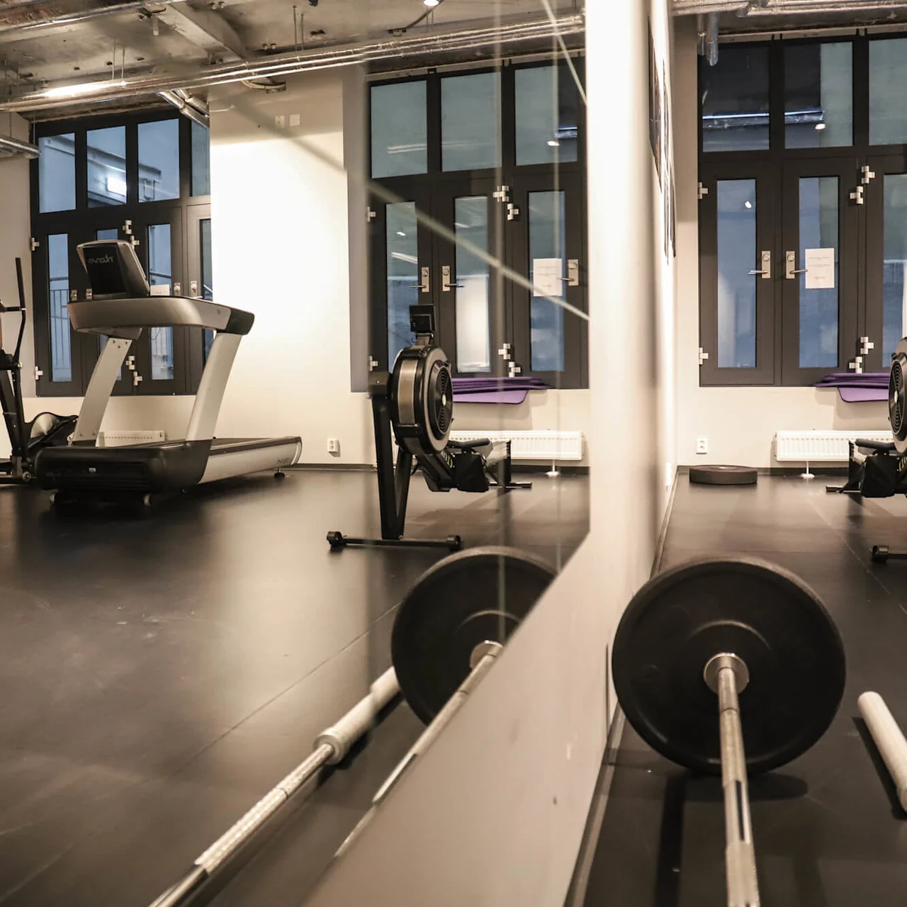 Löpband och roddmaskin står stilla i ett modernt gym med svart golv, stora fönster och exponerade takrör. En skivstång ligger på golvet, speglad i speglar.