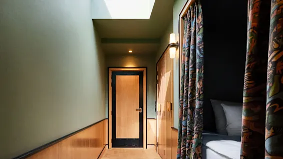 Trang hotellkorridor med mønstrede gardiner, tregulv og overlys, fører til en dør.