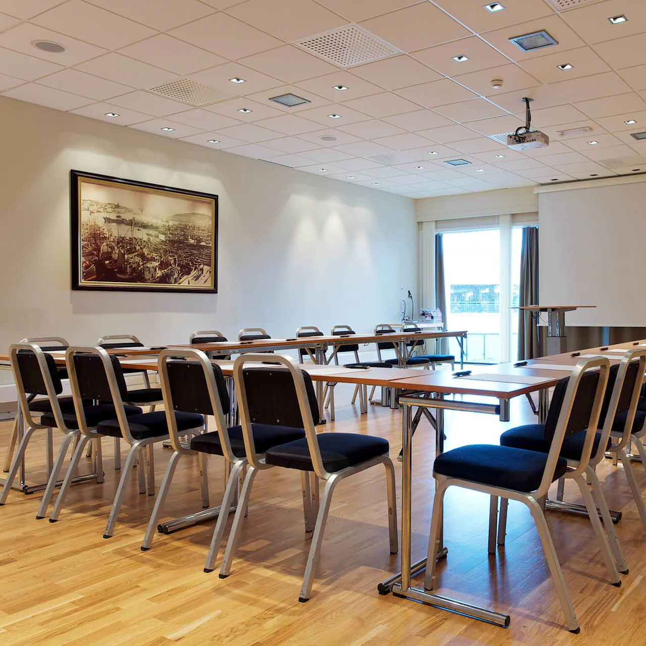 Konferanserom med rader av stoler og bord vendt mot en tom projektorskjerm. Et stort historisk maleri, klokke og potteplante pryder de kremfargede veggene; tregulv gir varme.