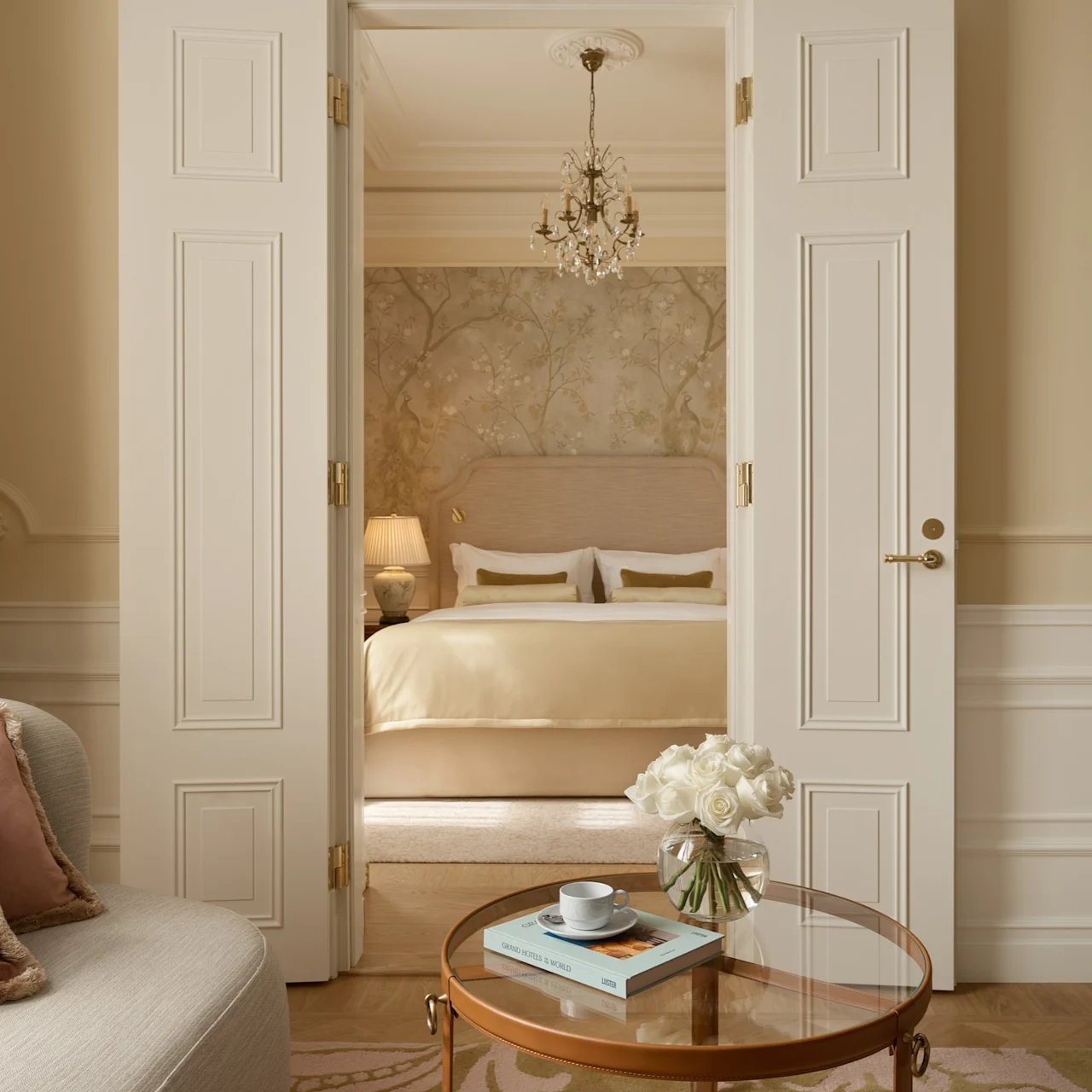 A coffee cup sits on a glass table with a book and roses. It faces a cozy bed beneath a chandelier, viewed through double doors in an elegant beige room.