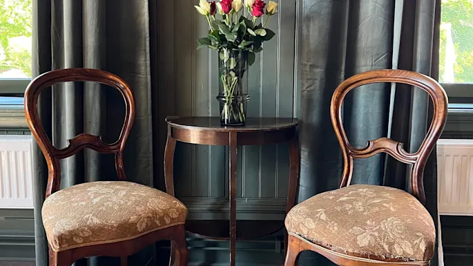 Et trebord har en vase med røde og hvite roser, flankert av to vintage stoler mot en bakgrunn av blomstrete tapet og gardiner.