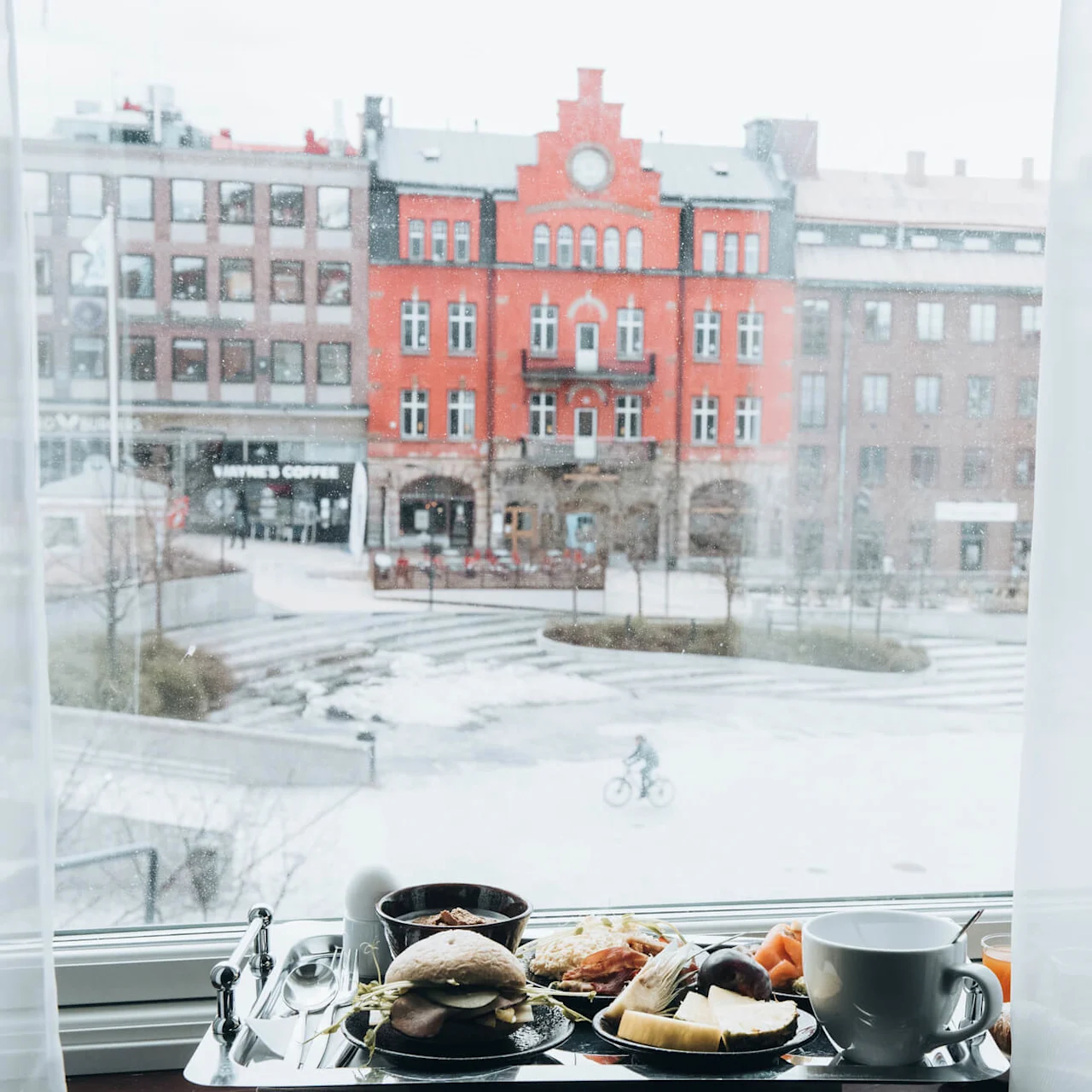 En frukostbricka med kaffe, smörgås och frukt står på en fönsterbräda och blickar ut över ett snötäckt stadsrum med en cyklist och byggnader, inklusive "Wayne's Coffee".