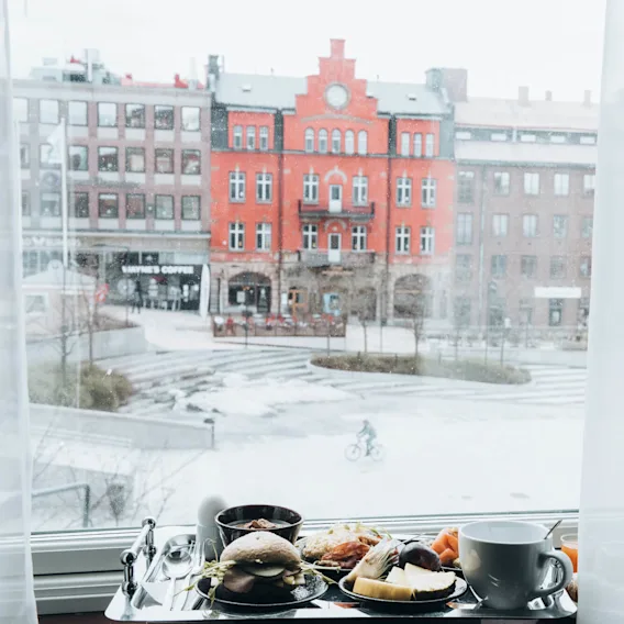 En frukostbricka med kaffe, smörgås och frukt står på en fönsterbräda och blickar ut över ett snötäckt stadsrum med en cyklist och byggnader, inklusive "Wayne's Coffee".