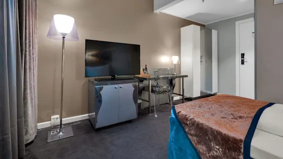 Et fjernsyn står på et skab og vender mod en seng med et brunt og blåt tæppe i et hotelværelse oplyst af gulv- og bordlamper.